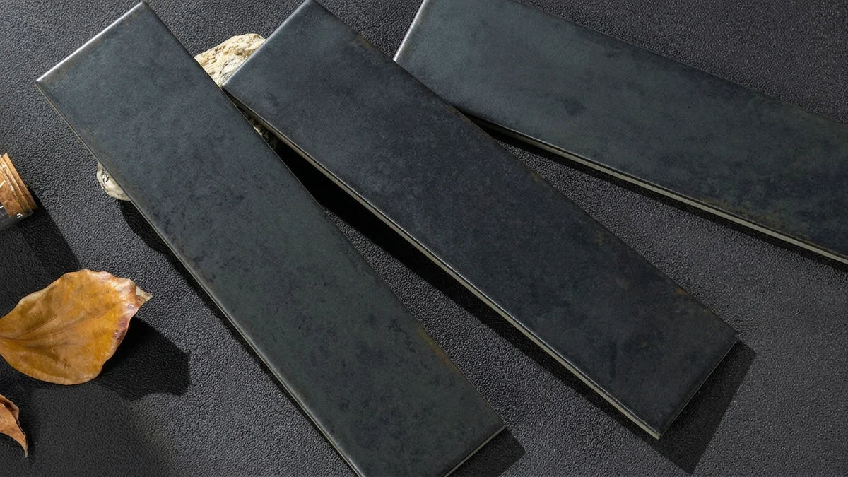 Three long rectangular dark charcoal gray stone slabs with a textured matte finish, arranged diagonally on a dark textured surface with a dried leaf and small decorative stones as props.