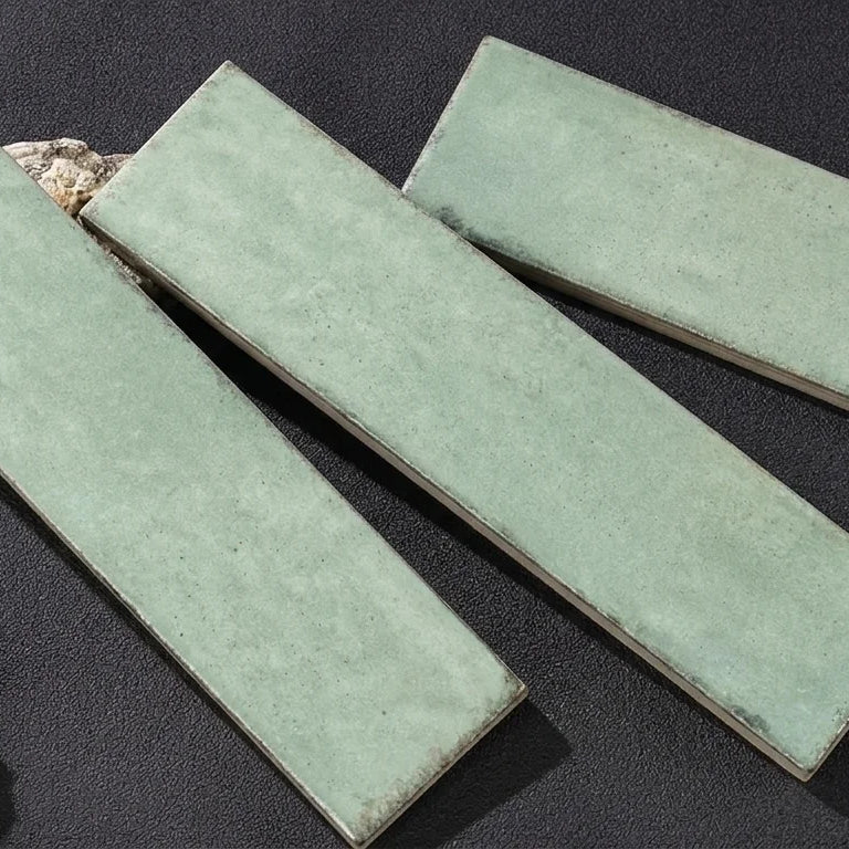 Three rectangular ceramic tiles with a matte sage green glaze, arranged diagonally on a dark textured surface with small decorative props.