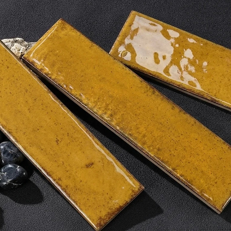 Three rectangular glossy ceramic tiles in a mustard yellow glaze with brown speckles, arranged diagonally on a dark textured surface with small decorative props.