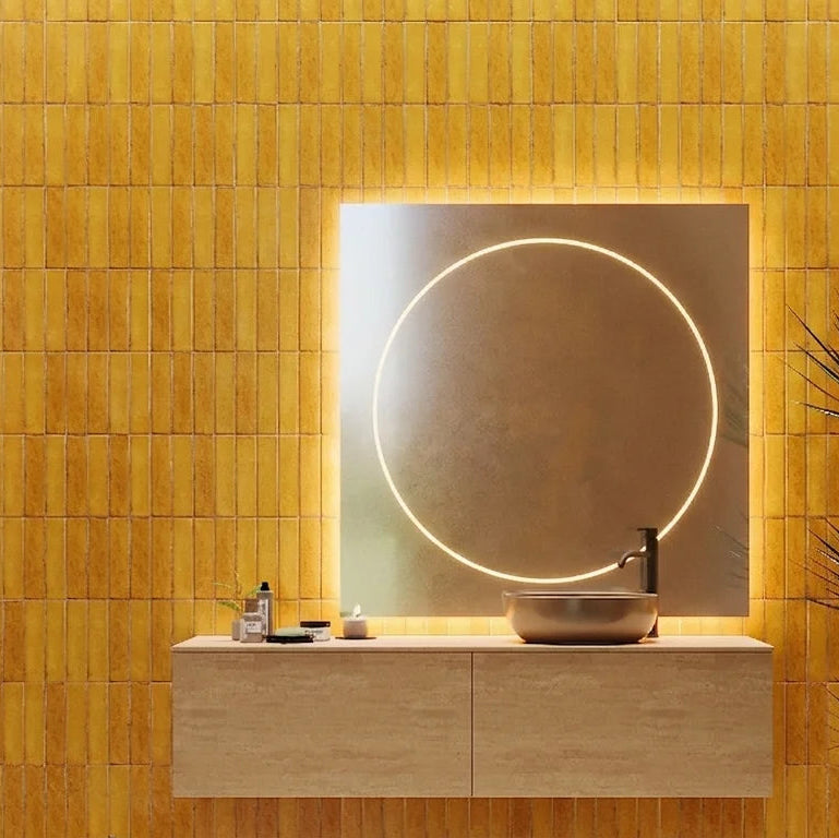 A modern bathroom vanity setup with a rectangular backlit mirror, a floating light wood console, a round vessel sink, and a tall dark plant against a warm yellow tiled wall.