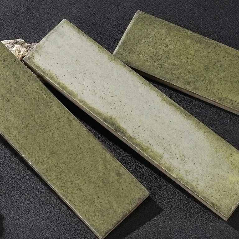 Three rectangular ceramic tiles with a textured sage green glaze, arranged diagonally on a dark textured surface with small decorative stones and a cork bottle.