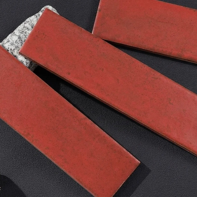 Three rectangular red ceramic subway tiles arranged diagonally on a dark textured surface, with a small decorative leaf and glass vial in the lower left corner.
