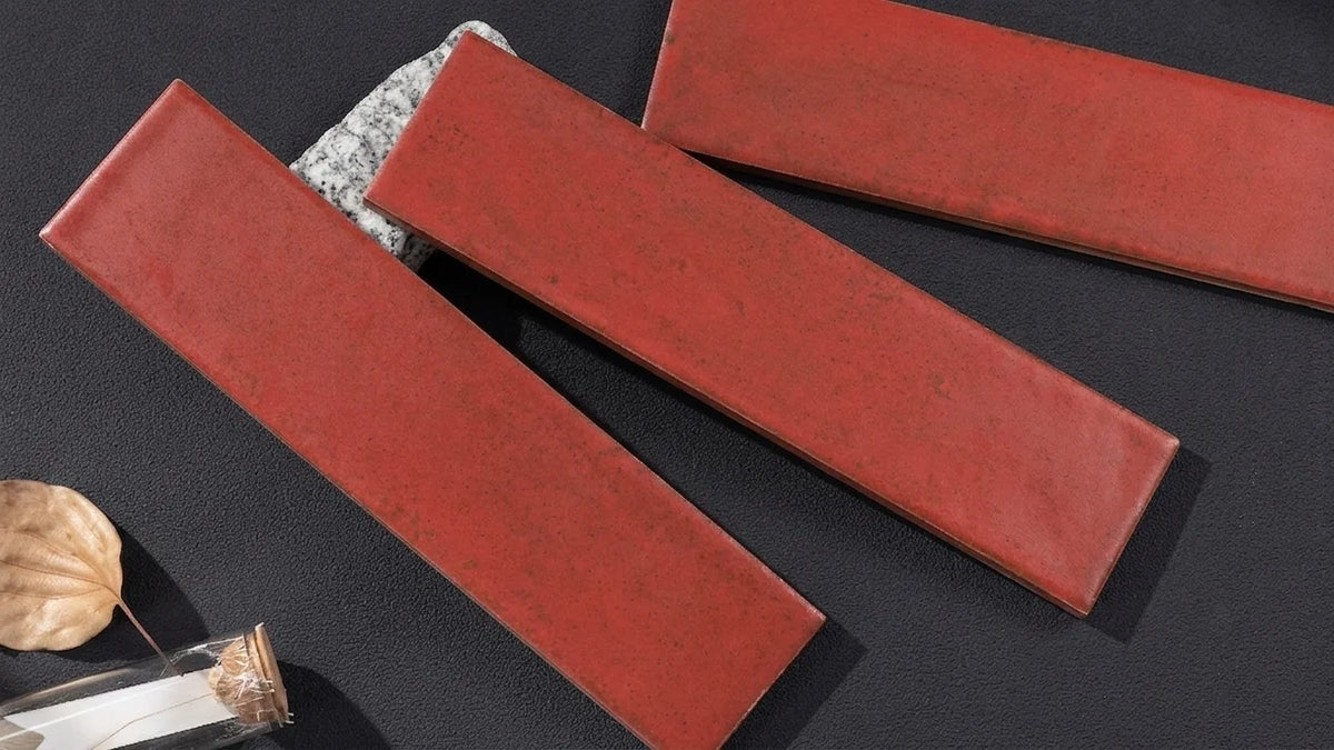 Three rectangular red ceramic subway tiles arranged diagonally on a dark textured surface, with a small decorative leaf and glass vial in the lower left corner.
