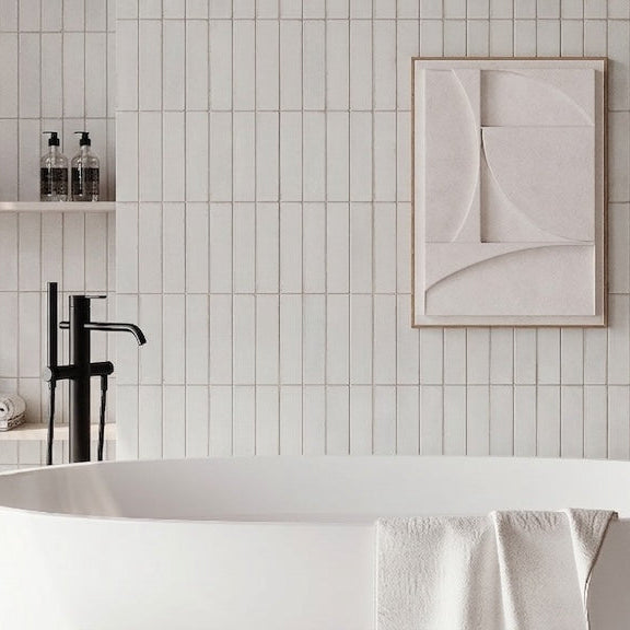 A modern freestanding white bathtub in a minimalist bathroom with white subway tiles, a black faucet, floating shelves with toiletries, and abstract wall art.