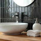 A white ceramic vessel sink with a chrome faucet, set on a light wood vanity in a bathroom with glossy black subway tiles, a white towel, and a soap dispenser.