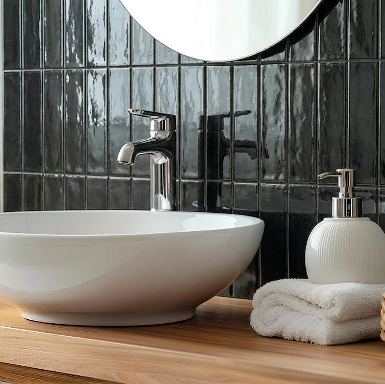 A white ceramic vessel sink with a chrome faucet, set on a light wood vanity in a bathroom with glossy black subway tiles, a white towel, and a soap dispenser.
