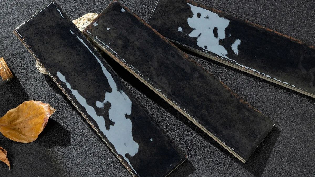 Three rectangular glossy black ceramic tiles with white marbled glaze accents, arranged diagonally on a dark textured surface with a dried leaf prop.