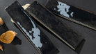 Three rectangular glossy black ceramic tiles with white marbled glaze accents, arranged diagonally on a dark textured surface with a dried leaf prop.