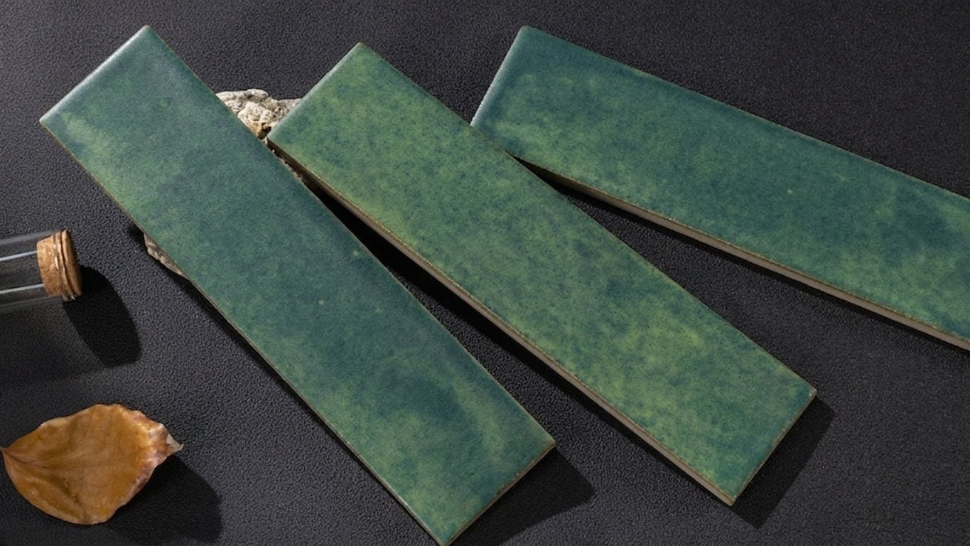 Three rectangular green ceramic tiles with a glossy glaze, arranged diagonally on a dark textured surface alongside a small cork stopper and a dried leaf.