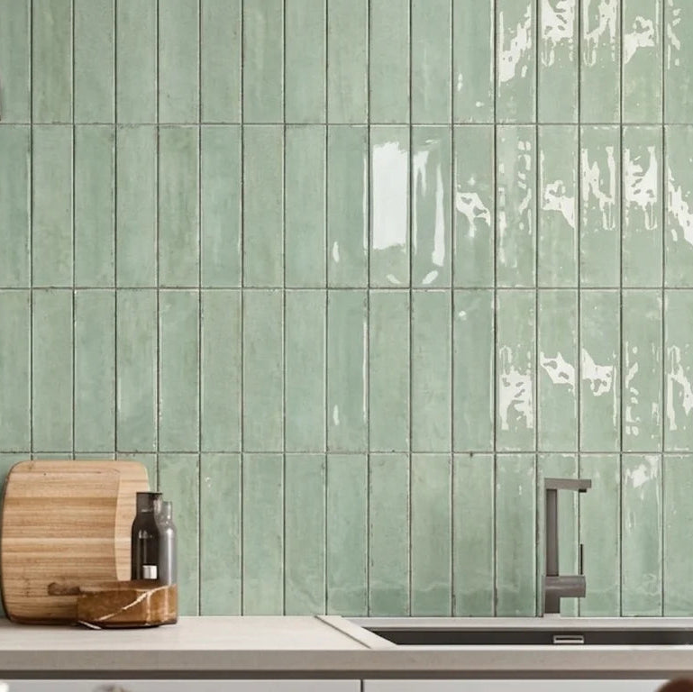 Green glossy rectangular subway tiles installed on a kitchen backsplash, with a stove, pot, wooden cutting board, and sink visible in the foreground.