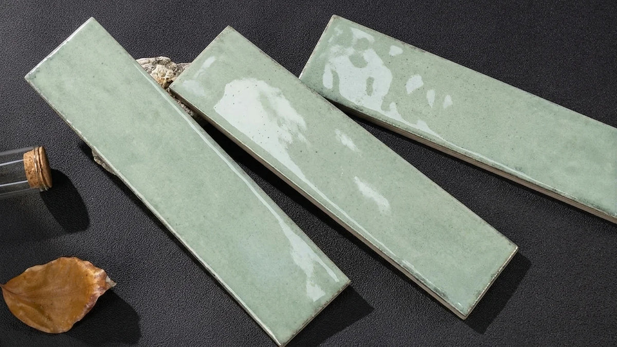 Three rectangular glossy ceramic tiles in a pale sage green glaze, arranged diagonally on a dark textured surface with small decorative props.