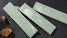 Three rectangular glossy ceramic tiles in a pale sage green glaze, arranged diagonally on a dark textured surface with small decorative props.