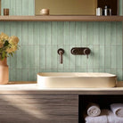 A modern bathroom vanity setup with a light wood countertop, a cream vessel sink, a green tiled backsplash, and a vase of cream flowers on the counter.
