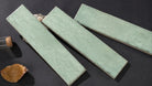 Three rectangular ceramic tiles with a matte sage green glaze, arranged diagonally on a dark textured surface with small decorative props.