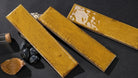 Three rectangular glossy ceramic tiles in a mustard yellow glaze with brown speckles, arranged diagonally on a dark textured surface with small decorative props.