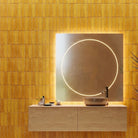 A modern bathroom vanity setup with a rectangular backlit mirror, a floating light wood console, a round vessel sink, and a tall dark plant against a warm yellow tiled wall.