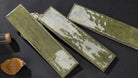 Three rectangular ceramic tiles with a distressed sage green and white glaze, arranged diagonally on a dark textured surface next to a cork stopper and a dried leaf.
