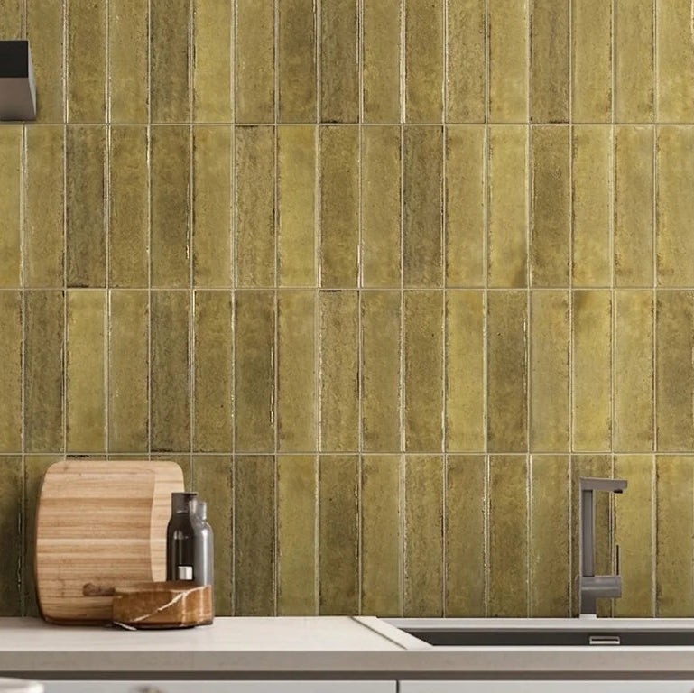 Vertical olive green subway tiles installed on a kitchen backsplash behind a stove, sink, and countertop with a cutting board and bottle.