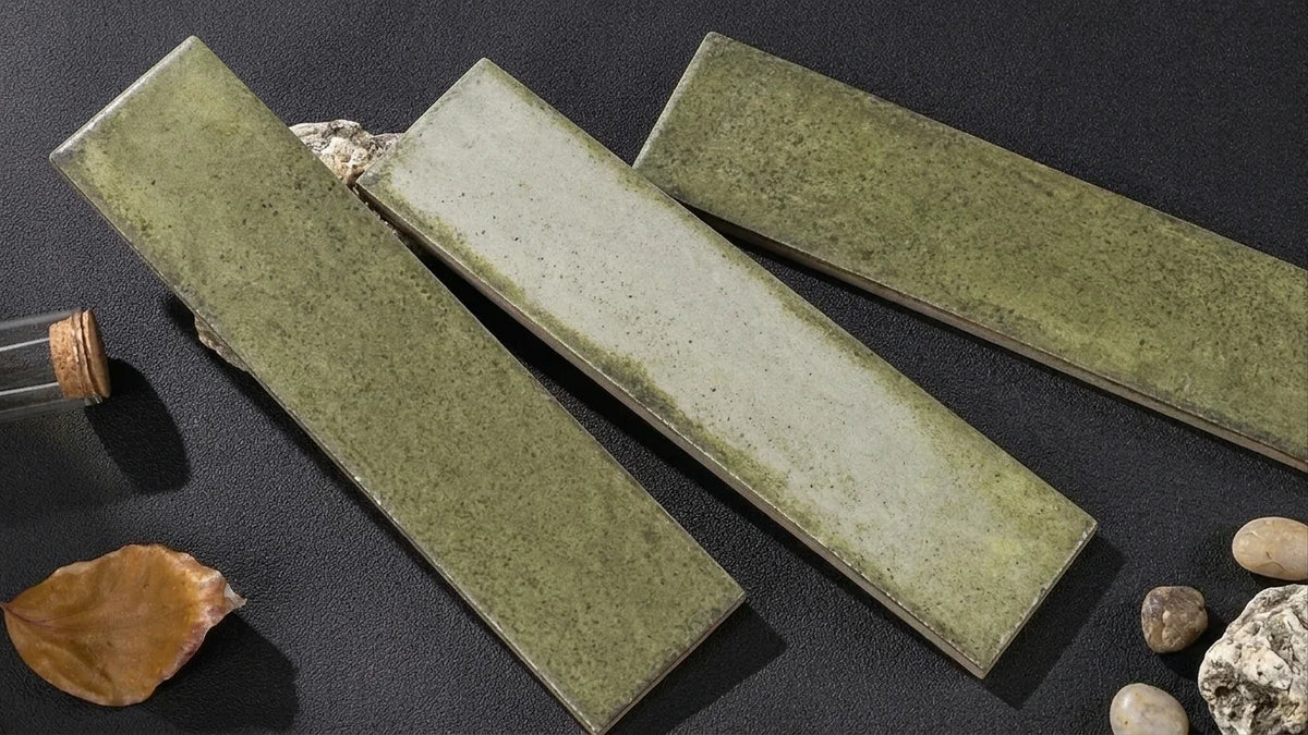 Three rectangular ceramic tiles with a textured sage green glaze, arranged diagonally on a dark textured surface with small decorative stones and a cork bottle.