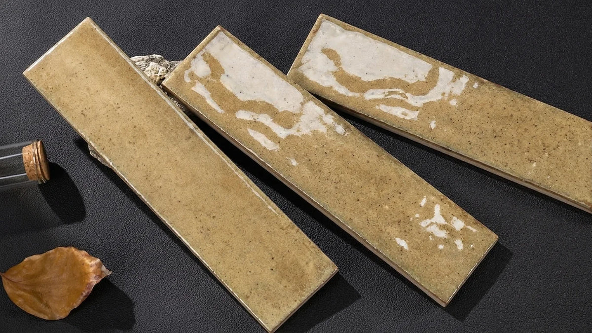 Three rectangular ceramic tiles with a textured beige and white marbled pattern, arranged diagonally on a dark textured surface next to a cork bottle and a dried leaf.