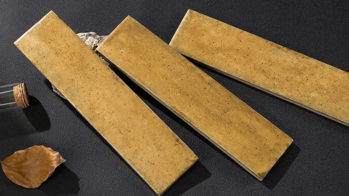 Three rectangular ceramic tiles with a textured, speckled ochre-brown glaze, arranged diagonally on a dark textured surface with a cork stopper and a dried leaf prop.