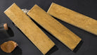 Three rectangular ceramic tiles with a textured, speckled ochre-brown glaze, arranged diagonally on a dark textured surface with a cork stopper and a dried leaf prop.