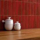 Red glossy rectangular wall tiles on a kitchen backsplash above a wooden countertop, with white ceramic containers and a potted green plant.