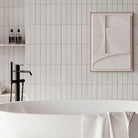 A modern freestanding white bathtub in a minimalist bathroom with white subway tiles, a black faucet, floating shelves with toiletries, and abstract wall art.